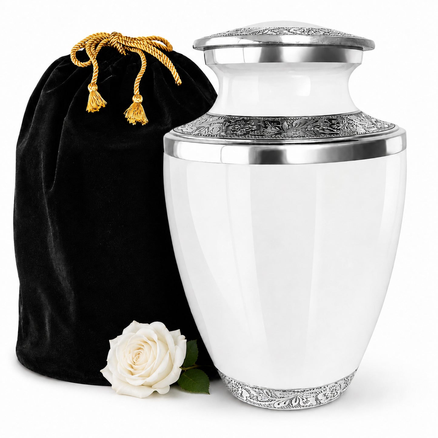 A white ceramic cremation urn with ornate silver metal detailing on the lid, rim, and base, shown next to a black velvet drawstring bag with gold tassel cords and a single white rose on a clean white background.